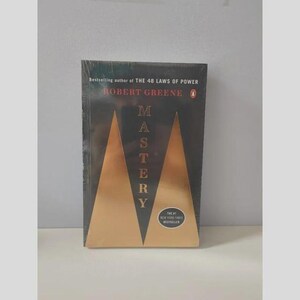 May include: A black book titled Mastery by Robert Greene, featuring a gold triangular graphic design on the cover. The text reads Bestselling author of THE 48 LAWS OF POWER, Robert Greene, Mastery, and The #1 New York Times Bestseller in a circular badge.