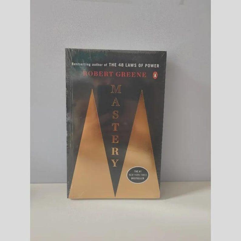 May include: A black book titled Mastery by Robert Greene, featuring a gold triangular graphic design on the cover. The text reads Bestselling author of THE 48 LAWS OF POWER, Robert Greene, Mastery, and The #1 New York Times Bestseller in a circular badge.