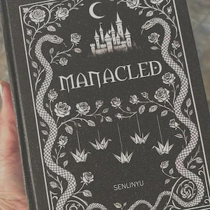 May include: A black book cover with white line art featuring a central castle silhouette under a crescent moon. The title MANACLED is printed in the center, surrounded by two large snakes, climbing rose vines, and three hanging paper cranes.