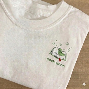May include: A white t-shirt featuring an embroidered design on the chest. The embroidery depicts a small green caterpillar wearing glasses, sitting on an open book with a red bookmark. The text "book worm" is stitched below in green thread.