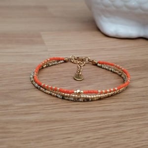 Peut inclure: Un bracelet multi-rangs composé de perles de rocaille orange vif, dorées métallisées et tachetées de blanc et de noir. Le bracelet comprend une chaînette d'extension en métal doré avec une petite breloque circulaire et un fermoir mousqueton.