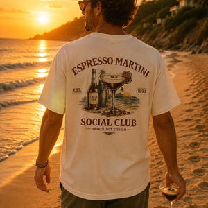 May include: An off-white t-shirt featuring a graphic on the back with a bottle, a cocktail glass, and the text "ESPRESSO MARTINI SOCIAL CLUB EST. 1993 SHAKEN, NOT STIRRED". The design includes a beach scene with palm trees and a sunset.