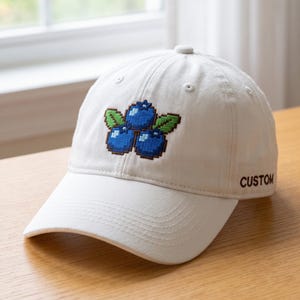 May include: A cream-colored baseball cap featuring an embroidered pixel art design of three blue blueberries with green leaves on the front panel and the word CUSTOM embroidered in dark brown thread on the side panel.
