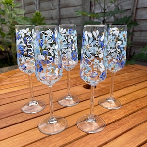 May include: Five clear glass champagne flutes featuring hand-painted blue and white floral designs. Each glass is personalised with a name in white script, including Claire, Annabel, Harriet, and Ellie, along with titles like Bride and Maid of Honour.