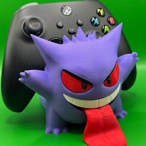 Può includere: Una statuetta del personaggio Gengar che funge da supporto per controller, con occhi rossi, un ampio sorriso bianco e una lunga lingua rossa che pende verso il basso, posizionata davanti a un controller di gioco wireless nero con tasti colorati.