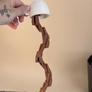Pode incluir: Uma chávena de café de cerâmica branca numa posição inclinada, com uma escultura sólida de cor castanha, ondulada e semelhante a um líquido, que flui da chávena para um pires branco a condizer numa superfície cinzenta texturada.