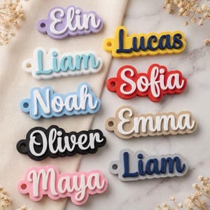 Puede incluir: Una colección de llaveros con nombres personalizados en varios colores, incluyendo lavanda, azul claro, blanco, negro, rosa, amarillo, rojo, beige y gris. Cada llavero presenta un nombre en relieve con un pequeño orificio circular.