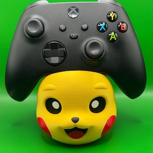Könnte beinhalten: Ein schwarzer kabelloser Videospiel-Controller, der auf einem gelben, 3D-gedruckten Charakterkopf-Ständer ruht. Der Controller verfügt über ein Steuerkreuz, zwei Analogsticks und vier farbige Aktionstasten mit den Beschriftungen X, Y, A und B.
