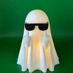 Könnte beinhalten: Eine weiße, geisterförmige Figur mit strukturierter Oberfläche, die eine schwarze Sonnenbrille trägt. Die Figur hat zwei erhobene Arme, die jeweils den Mittelfinger zeigen. Ein warmes, sanftes gelbes Licht leuchtet aus dem Inneren des Geistes.