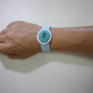 Può includere: Un braccialetto azzurro con un componente centrale ovale color verde acqua con un bordo bianco. Il componente verde acqua presenta le lettere EM stampate in nero. Il cinturino è realizzato in tessuto a coste e si chiude con una chiusura piatta.