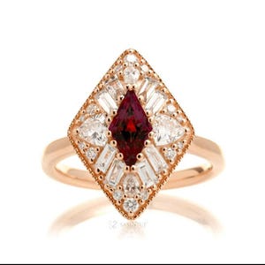 May include: A rose gold ring featuring a central marquise-cut deep red gemstone surrounded by a geometric halo of baguette and round-cut white diamonds. The setting is shaped like a diamond, with milgrain detailing along the outer edges.