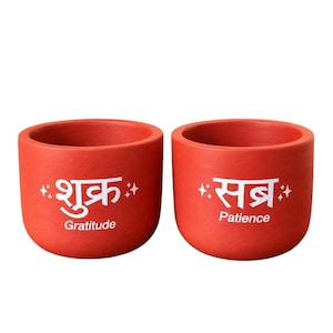 May include: Two cylindrical red terracotta pots featuring white text and star accents. The left pot displays the Hindi word "शुक्र" above the English word "Gratitude." The right pot displays the Hindi word "सब्र" above the English word "Patience."