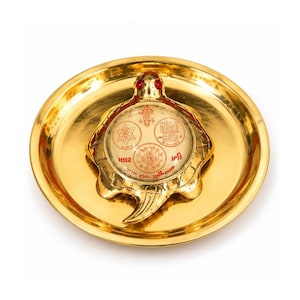 Puede incluir: Una figura de tortuga de metal dorado con dos ojos de piedra roja, colocada sobre un plato redondo de metal dorado a juego. El caparazón de la tortuga presenta una inserción de vidrio circular con símbolos geométricos y texto en rojo.