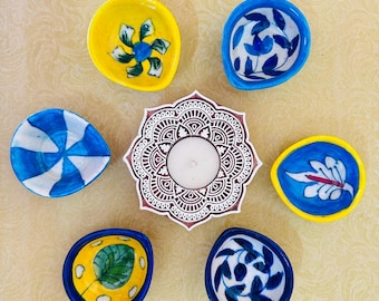 May include: A central white candle holder with a brown mandala-style carved pattern surrounded by six small, hand-painted ceramic bowls. The bowls feature various blue, yellow, and white floral or geometric patterns on their interior surfaces.