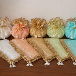 Pode incluir: Cinco bolsas com cordão e cinco panos retangulares a condizer em branco, pêssego, dourado, verde-claro e turquesa. Cada item apresenta um tecido transparente, borda de renda metálica dourada e contas de pérolas brancas nos cordões e cantos.