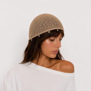 Può includere: Una calotta all'uncinetto color beige caratterizzata da un motivo a griglia a trama aperta e da una serie di piccoli ciondoli sferici in metallo color argento che pendono lungo il bordo inferiore, creando un copricapo leggero e strutturato.