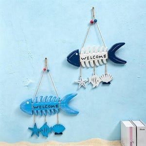 Op de afbeelding: Twee houten wandhangers in de vorm van visgraten met het woord WELCOME in het midden. De ene is donkerblauw en wit, de andere is lichtblauw. Beide hebben hangende ornamenten van een zeester, een kleine vis en een schelp aan touw.