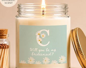 Op de afbeelding: Een brandende kaars in een glazen pot met een lichtblauw etiket met een witte letter C, madeliefjes en de tekst Will you be my bridesmaid? Een klein glazen flesje met een kurk gevuld met houten lucifers staat ernaast.