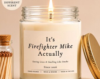 Op de afbeelding: Een glazen kaars in een pot met een brandende lont en een wit etiket met de tekst It's Firefighter Mike Actually, Saving Lives & Smelling Like Smoke, Since 2026. Daarnaast staat een klein glazen flesje met lucifers en een gouden metalen deksel.