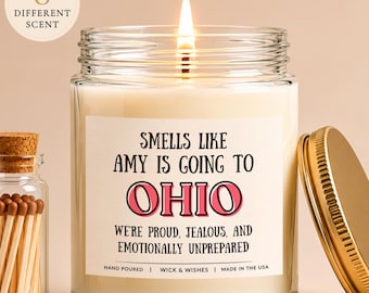 Op de afbeelding: Een doorzichtige glazen kaarsenpot met een brandende lont en een wit etiket met de tekst "SMELLS LIKE AMY IS GOING TO OHIO WE'RE PROUD, JEALOUS, AND EMOTIONALLY UNPREPARED". Daarnaast staat een klein glazen flesje gevuld met lucifers.