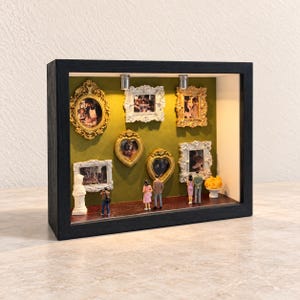 May include: A black shadow box frame containing a miniature art gallery with a green wall, a dark floor, and two overhead lights. The display features six ornate gold and silver picture frames, a white bust, and four small human figurines.