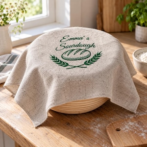 May include: A beige linen bread proofing cover draped over a round banneton basket. The fabric features dark green embroidery with the text Emma's Sourdough, a central illustration of a loaf of bread, and two wheat stalks framing the bottom.