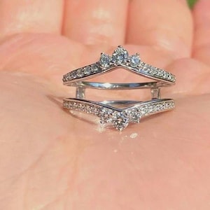 May include: A silver-toned ring guard featuring two chevron-shaped bands connected by vertical bars. The bands are encrusted with small, clear, round-cut stones, with a larger, prominent stone set at the center point of each chevron.