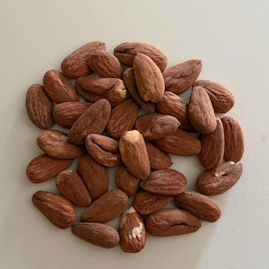 Pode incluir: Um grupo de amêndoas inteiras e cruas com casca castanha e texturizada. Cada amêndoa tem uma forma alongada de gota com estrias visíveis e variações naturais de cor, variando do bege claro ao castanho avermelhado profundo.