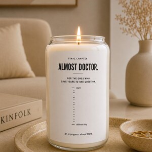 May include: A white wax candle in a clear glass jar with a lit wick. The label reads: FINAL CHAPTER ALMOST DOCTOR. FOR THE ONES WHO GAVE YEARS TO ONE QUESTION. A progress bar spans from start to defense day. Text at the bottom reads: dr. in progress. almost there.