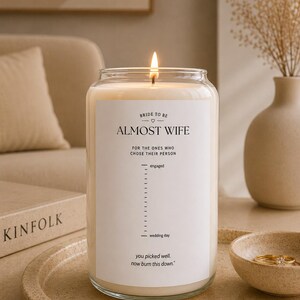 Könnte beinhalten: Eine brennende Kerze in einem klaren Glasgefäß mit einem weißen Etikett, das den Text BRIDE TO BE ALMOST WIFE FOR THE ONES WHO CHOSE THEIR PERSON, eine Countdown-Skala von engaged bis wedding day und den Text you picked well. now burn this down. enthält.