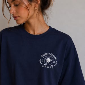 Könnte beinhalten: Ein dunkelblaues T-Shirt mit kurzen Ärmeln und einer weißen kreisförmigen Grafik auf der Brust. Das Design enthält den Text "GARRETT GRAHAM", "HAWAII UNIVERSITY", "NO 44", "OFF CAMPUS" und "HAWKS" um eine zentrale Abbildung eines Helms und gekreuzter Stöcke.