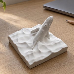 May include: A white sculpture featuring a whale emerging from a textured, wavy base that mimics the appearance of ocean water. The whale is positioned at an angle, with its body partially submerged in the sculpted white surface.