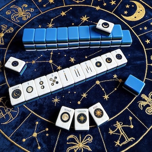 May include: A set of Mahjong tiles featuring celestial designs with moons, stars, and constellations. The tiles have white faces with blue, green, and dark blue layered backs. Some tiles display numbers, the word JOKER, or SOAP, GREEN, and RED.