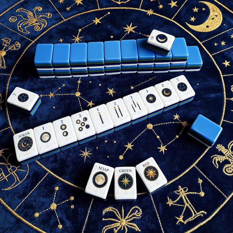 May include: A set of Mahjong tiles featuring celestial designs with moons, stars, and constellations. The tiles have white faces with blue, green, and dark blue layered backs. Some tiles display numbers, the word JOKER, or SOAP, GREEN, and RED.