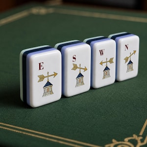 May include: Four rectangular mahjong tiles with white faces and layered blue and light blue sides. Each tile features a red letter E, S, W, or N above a graphic illustration of a gold weather vane with an arrow pointing in different directions.