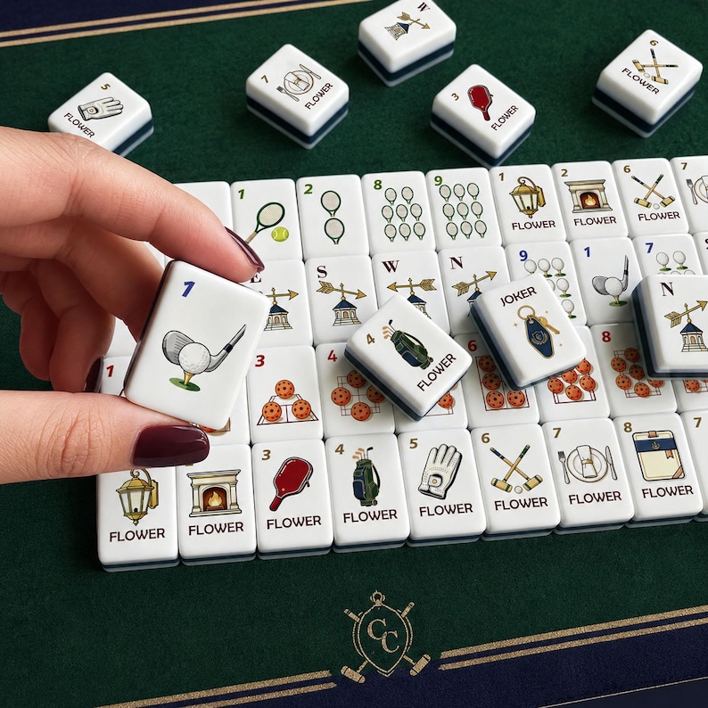 May include: A set of white Mahjong tiles featuring colorful illustrations of golf equipment, sports gear, and various icons. Each tile displays a number or letter, with some labeled as FLOWER or JOKER. The tiles are arranged on a dark green surface.