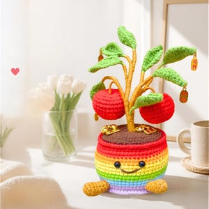 以下が含まれることがあります： 笑顔が刺繍された虹色の編みぐるみの鉢植え。緑の葉、2つの赤い丸い果実、枝からぶら下がった金色のチャームが付いた小さな木が入っています。鉢には2つの小さなベージュの足があり、上部には茶色の土が表現されています。
