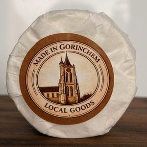 Op de afbeelding: Een rond, in wit papier verpakt pakket met een rond bruin etiket. Het etiket toont een illustratie van een kerktoren en de tekst "MADE IN GORINCHEM" bovenaan en "LOCAL GOODS" onderaan, gecentreerd op de papieren verpakking.