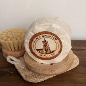 Op de afbeelding: Een rond, in wit papier verpakt item met een rond etiket met een illustratie van een kerk en de tekst MADE IN GORINCHEM LOCAL GOODS. Het staat op een ronde houten basis naast een ronde houten borstel met lichtgekleurde haren en een loofah.