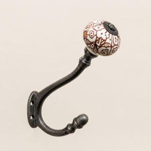 May include: A wall-mounted coat hook featuring a dark, weathered metal finish with a curved design. The tip of the hook is topped with a round, white ceramic knob decorated with a brown floral pattern and a small orange accent dot.