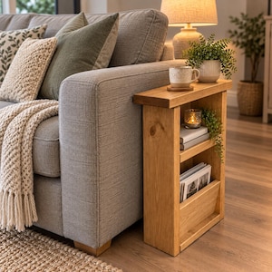 May include: A narrow, rectangular wooden side table with a light brown finish, featuring a flat top surface and two open storage shelves. The table is positioned beside a grey sofa and holds a ceramic mug, a small candle, and several magazines.