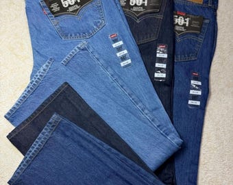 May include: Three pairs of Levi's 501 Original jeans in light blue, dark indigo, and medium blue denim. Each pair features a tan leather patch on the back waistband, a black 501 tag on the back pocket, and multiple paper tags attached to the fabric.