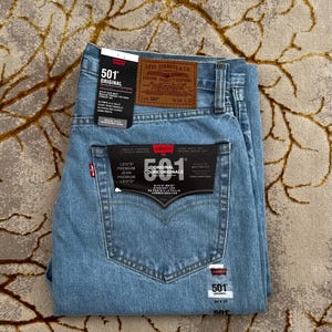 以下が含まれることがあります： ボタンフライ仕様のライトブルーのLevi's 501オリジナルストレートレッグジーンズ。ウエスト背面にW34 L32と記された茶色のレザーパッチがあり、501オリジナルのフィット感を説明する複数の白黒の製品タグが付いています。