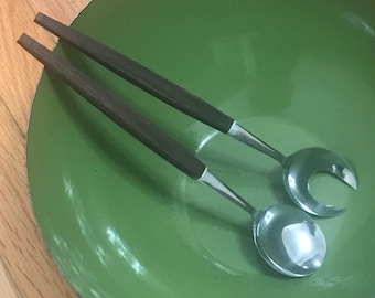 May include: A set of two salad servers with wooden handles. The servers are made of stainless steel and have a polished finish. The servers are sitting in a green bowl.
