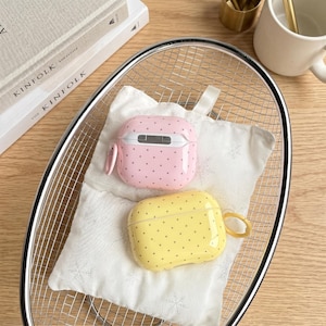 May include: Two protective cases for wireless earbuds, one in light pink and one in light yellow, both featuring a pattern of small dark dots. Each case includes a small circular ring attachment on the side for hanging or securing the device.