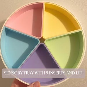 Peut inclure: Un plateau sensoriel circulaire comprenant cinq compartiments en forme de parts, aux couleurs pastel rose, jaune, vert clair, violet et bleu. Le plateau possède un cadre extérieur blanc et un couvercle. Le texte SENSORY TRAY WITH 5 INSERTS AND LID est visible.