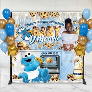 May include: A backdrop banner featuring a blue brick wall, a blue oven, and a blue monster character. The text reads There's a cookie in the oven and D'Mourie. The design includes chocolate chip cookie graphics, white milk drips, and gold and blue balloons.