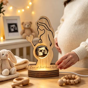 Könnte beinhalten: Eine hellfarbene Holzlampe in Form einer schwangeren Silhouette mit einem kreisförmigen, beleuchteten Zentrum, das ein Ultraschallbild zeigt. Der Sockel trägt den eingravierten Text Love at first heart beat. Die Lampe hat eine glatte Oberfläche.