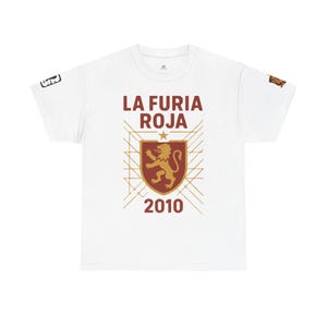May include: A white short-sleeve t-shirt featuring the text LA FURIA ROJA and 2010 in dark red. A central shield graphic displays a gold lion, topped with a star and surrounded by thin gold geometric lines. Small logos appear on both sleeves.