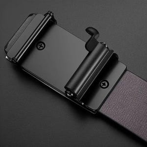May include: A matte black metal belt buckle with a rectangular frame and a sliding locking mechanism. The buckle features two visible Phillips head screws on the front plate and is attached to a dark brown textured belt strap with a visible grain.